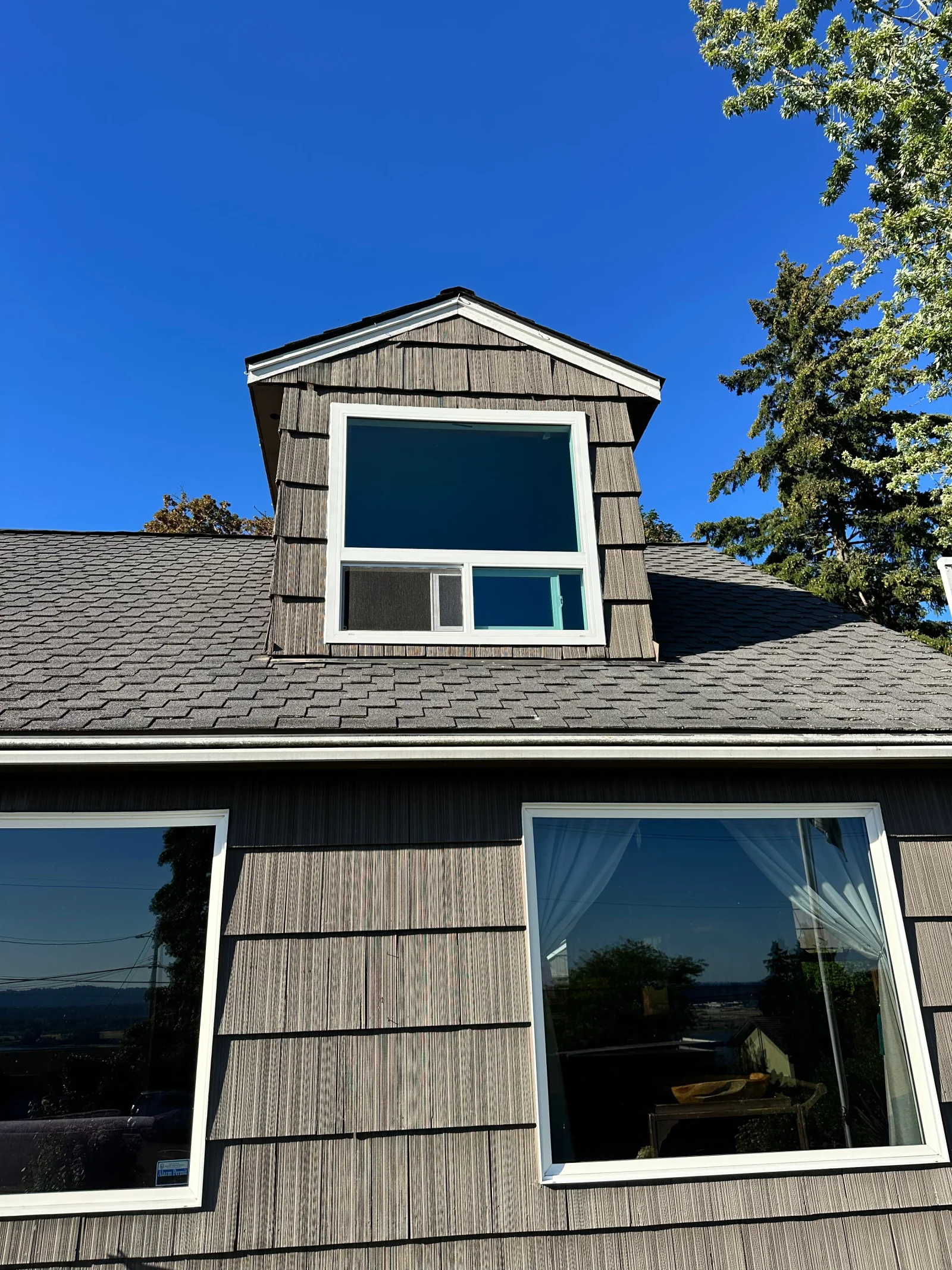 Dormer with white-framed window and large picture windows set in weathered wood shingle siding under asphalt shingle roof