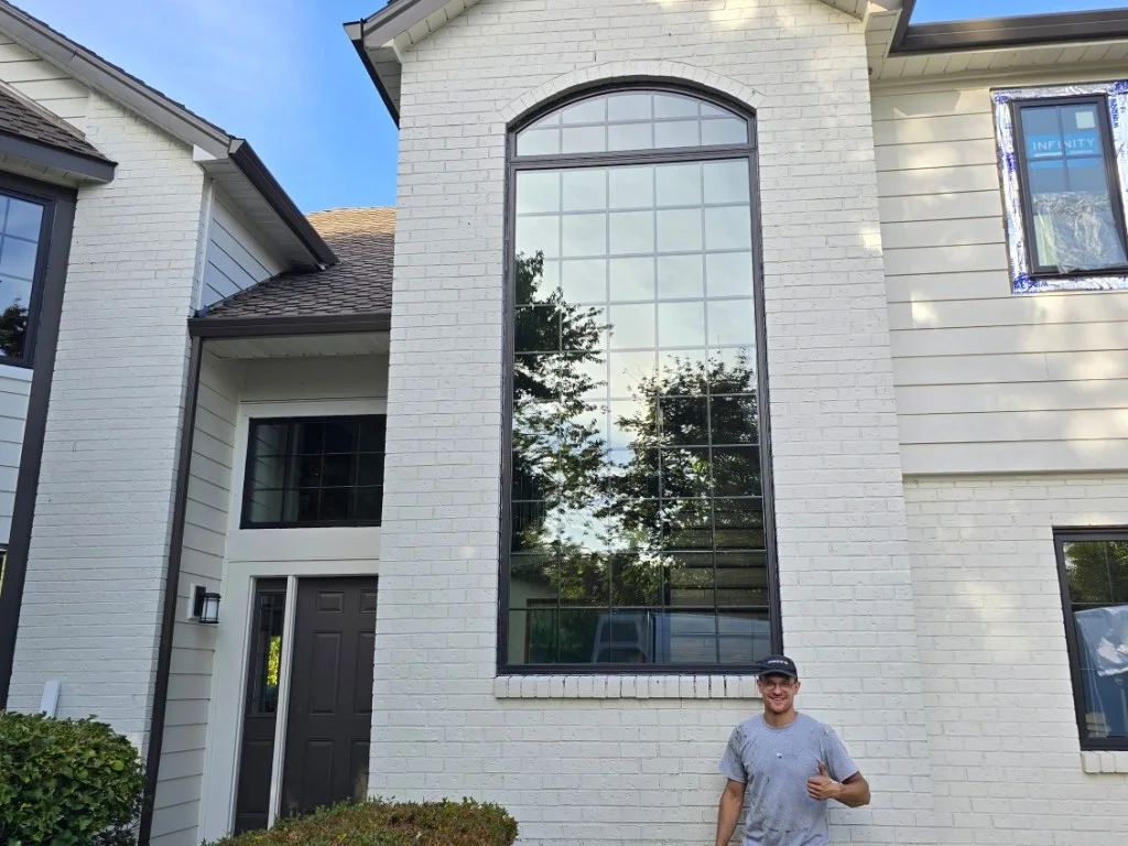 Tall arched grid picture window with dark bronze frame on white painted brick home with new window wrapped in protective film