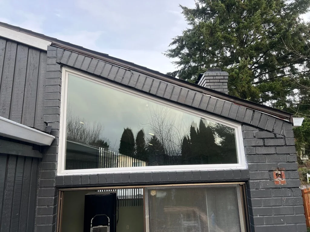 Large angled white-framed picture window set under a sloped roofline in dark gray brick and wood siding