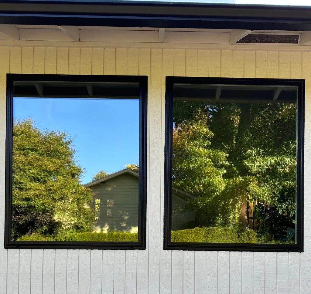 Two large black-framed picture windows set in cream vertical board siding with dark fascia above