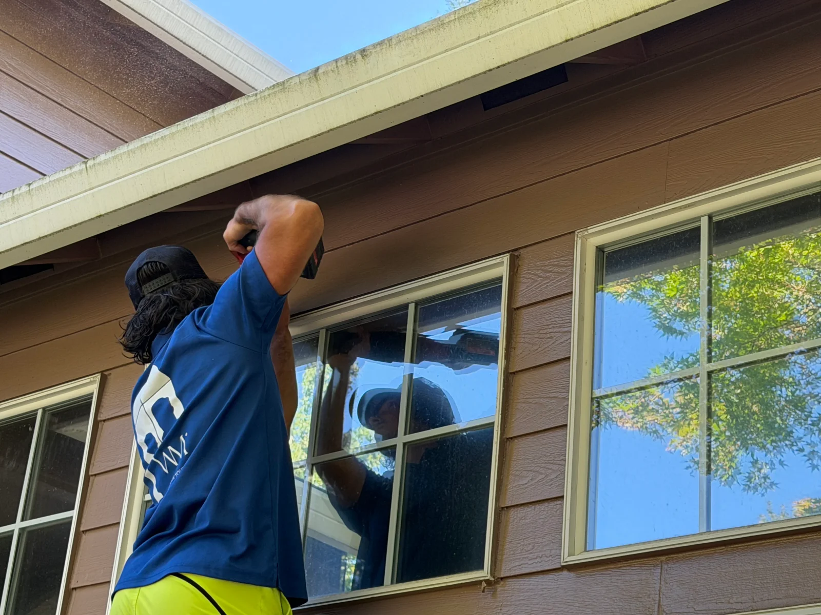Technician in blue shirt servicing a multi-pane window on brown lap siding house with cream trim
