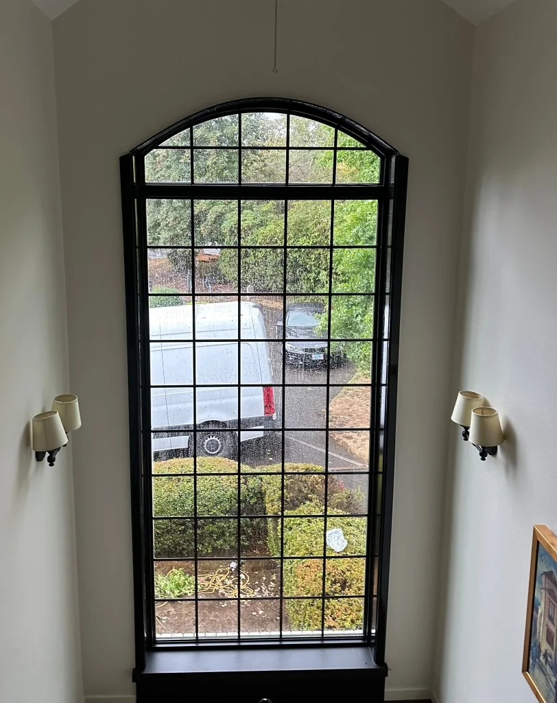Tall arched window with black frame and divided grid panes installed between white walls, with dark sill